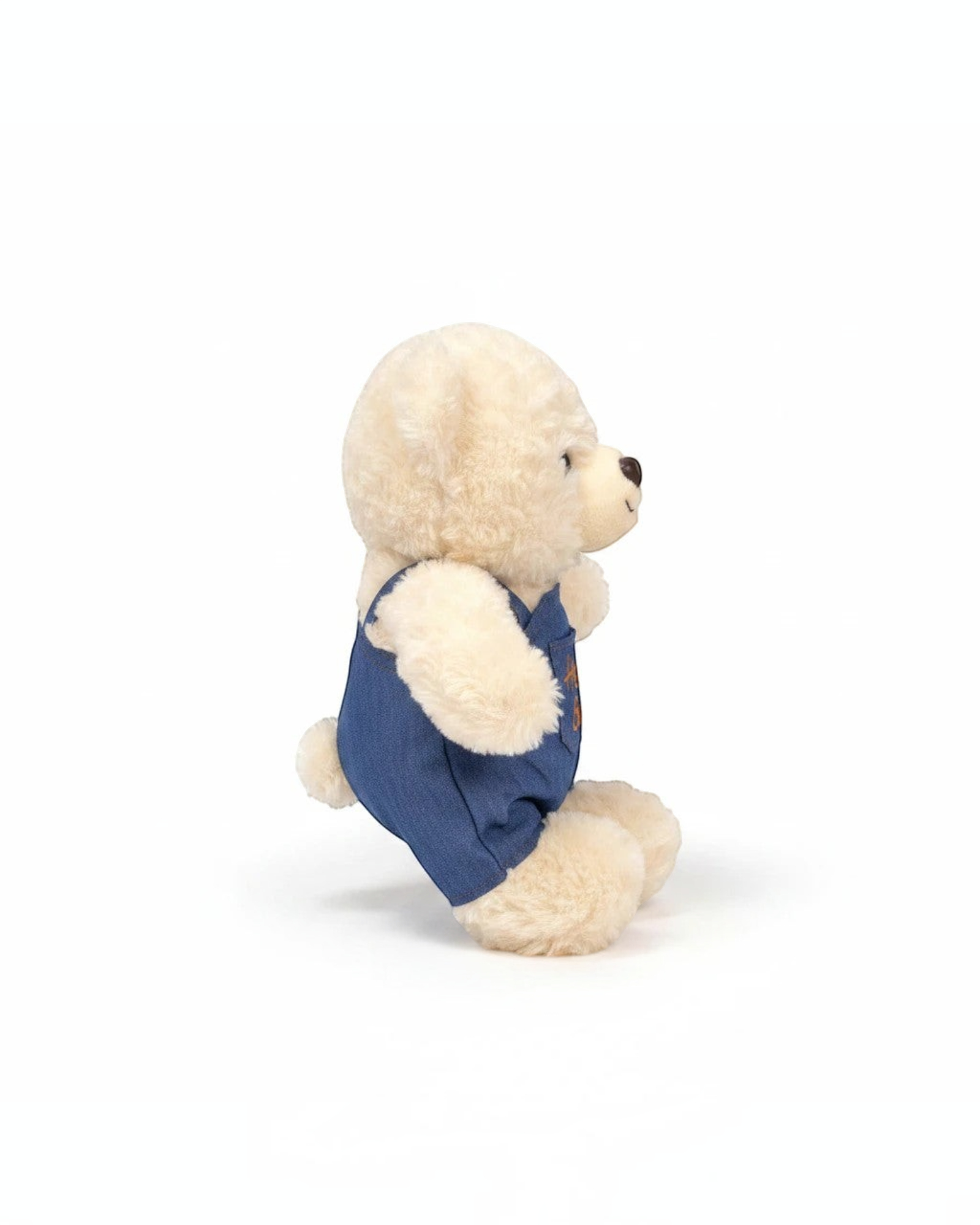 دب محشو ناعم من TagerlSaada بفرو كريمي يرتدي وزرة جينز زرقاء مطرزة بعبارة Hello Bear، منظر جانبي.